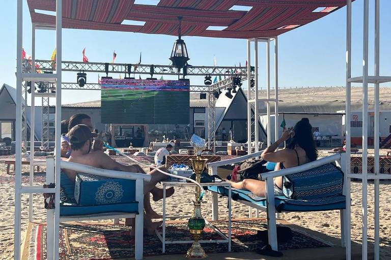 casal assiste partido no telão debaixo de tenda no deserto, com vista para o mar, em beach clube nos arredores de Doha