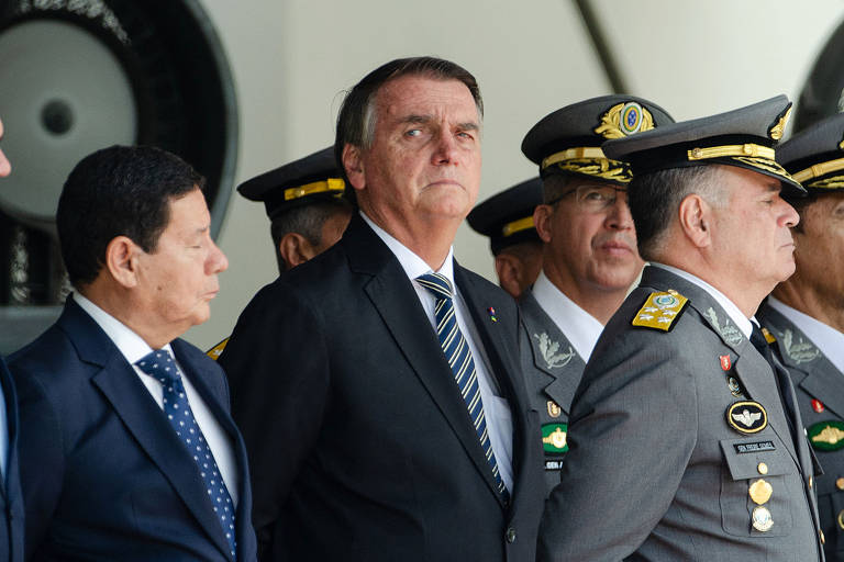 Da esquerda pra direita, Mourão de perfil, Bolsonaro, que está de frente olhando para a direita, ao lado dele. Ambos vestem terno e gravata, posicionados atrás de Freire Gomes, de farda cinza e quepe, com outros militares ao fundo