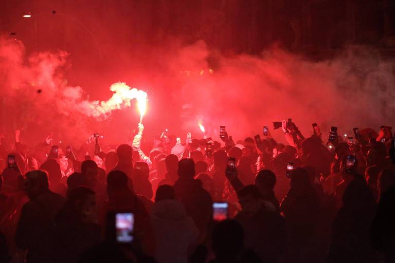 Imagem colorida mostra uma sombra com várias pessoas, com uma fumaça avermelhada, provocada por um sinalizador de fogos de artifício