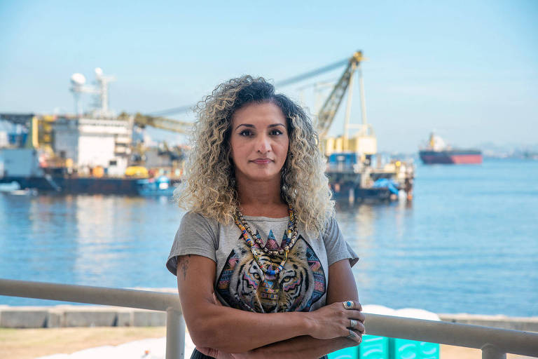 Bruna é uma mulher trans. Tem cabelo crespo, longo e loiro. Está em pé com os braços cruzados e séria. Ao fundo, desfocados, alguns navios no mar.