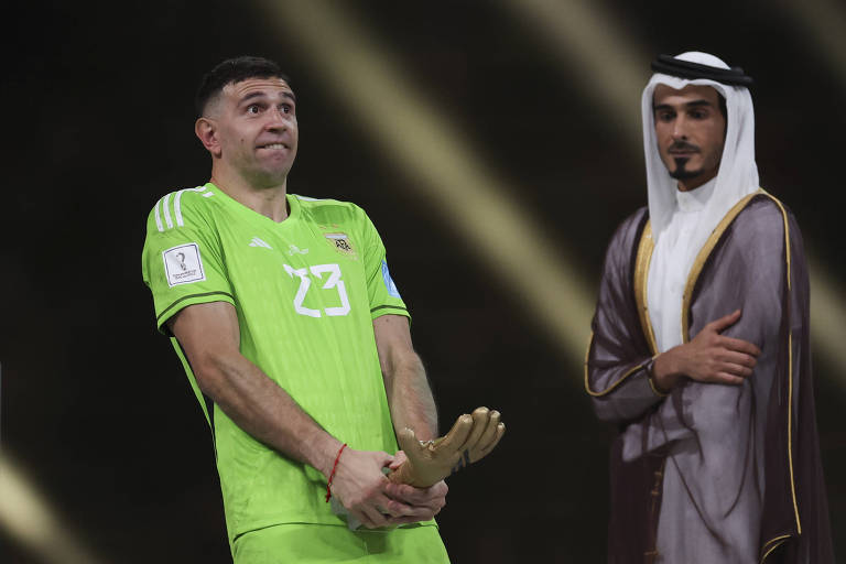 NO estádio Lusail, usando uniforme verde, o goleiro Emiliano Martínez, da Argentina, exibe abaixo do abdômen, em gesto obsceno, o prêmio Luva de Ouro, que é um troféu na forma de uma mão aberta, dado ao melhor goleiro da Copa do Qatar 
