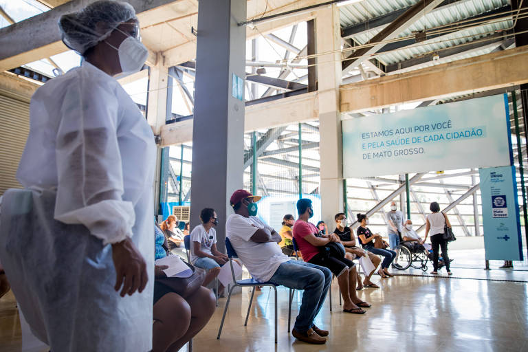 Imagem mostra uma mulher, profissional da saúde, em primeiro plano e ao fundo pessoas sentadas esperando no centro de triagem para atendimento de pacientes com coronavírus em Cuiabá