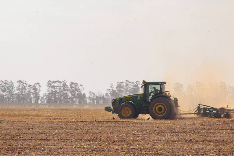 Máquina agrícola pilotada por uma pessoa passa em um terreno plano, sem plantação, apenas de terra