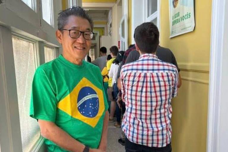 O empresário Winston Ling está usando uma camiseta da bandeira Brasil. Ele posa para a foto em um corredor, onde há outras pessoas de costas.