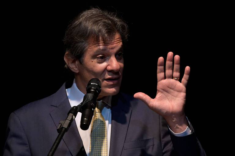 Fernando Haddad está com a mão levantada enquanto fala em frente a um microfone.