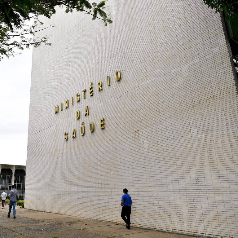 Fachada de pastilhas brancas onde se lê "Ministério da Saúde" em letras de forma douradas. Há pessoas andando em frente