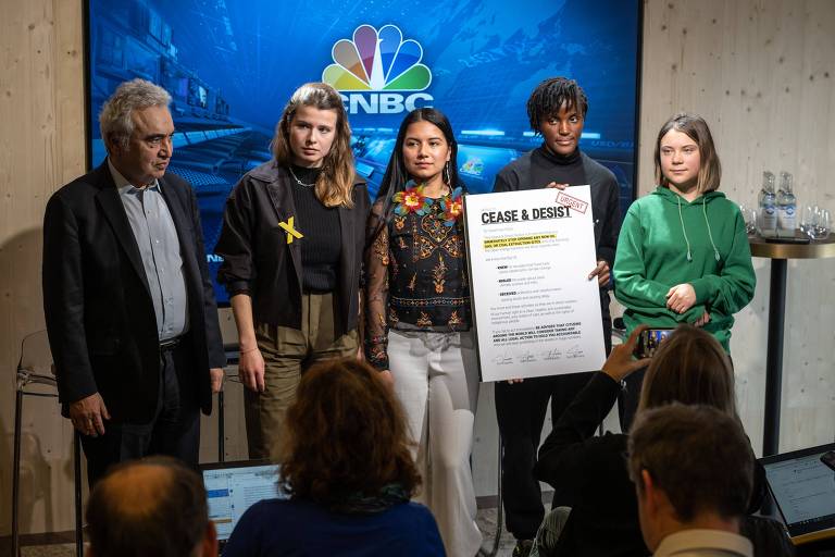 O grupo está de pé, posando para fotos, enquanto Vanessa segura um cartaz com cópia da carta