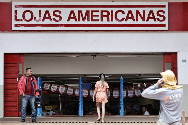 Fachada de uma loja da Americanas com a logomarca da rede acima da porta de entrada e 3 pessoas à frente, uma mulher ao centro, entrando na loja, um homem parado à esquerda e outro à direita