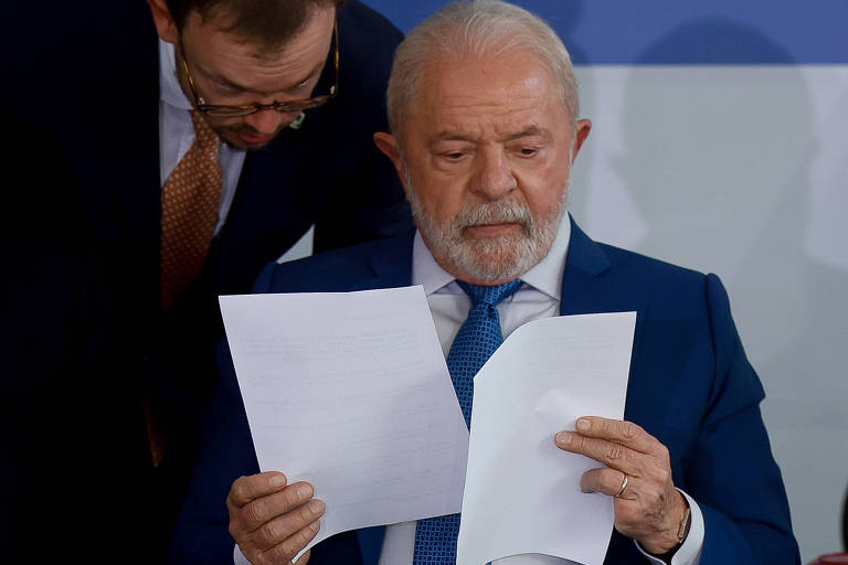 Sentado, segurando uma folha de papel branco em cada mão e com um homem branco, de óculos e terno escuro falando próximo ao seu ouvido direito, Lula, um homem branco, de cabelos e barba da mesma cor, usa um terno azul com gravata da mesma cor e camisa branca