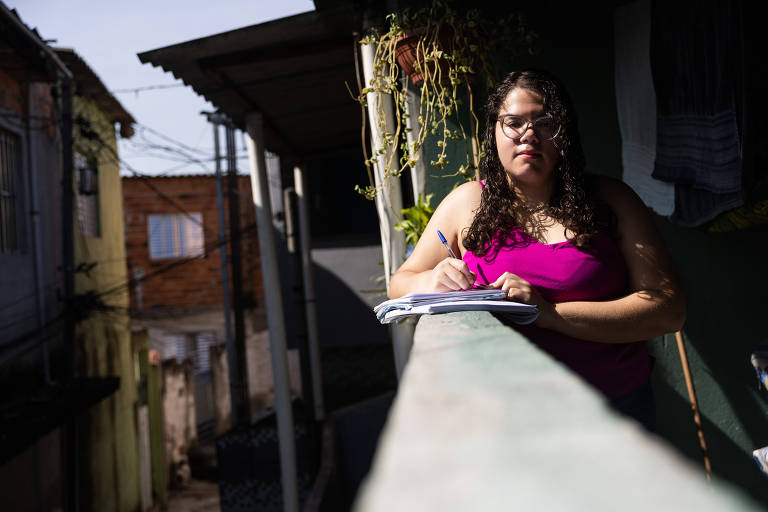 Estudante escreve em caderno na sacada de sua casa; ao fundo aparecem barracos