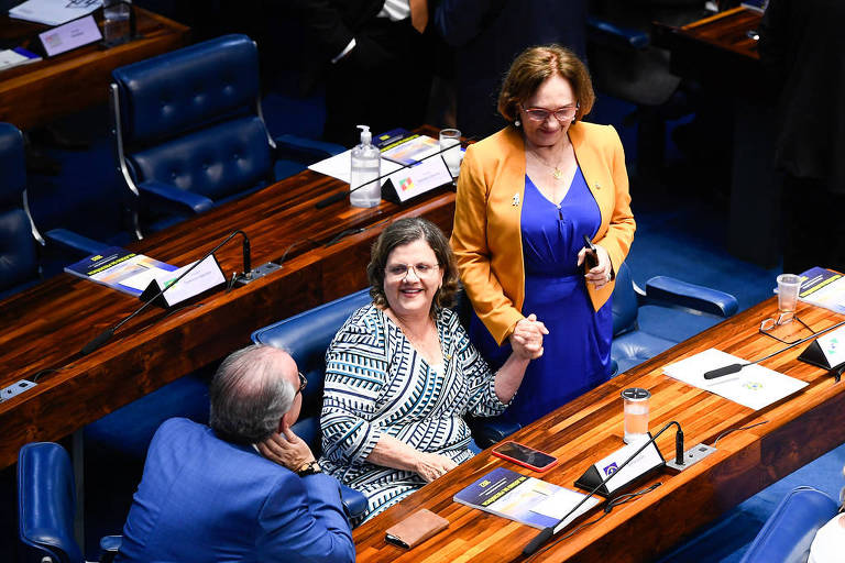 A senadora Teresa Leitão está sentada no plenário do Senado conversando com um homem, que está de costas. Ela está de mãos dadas com a senadora Zenaide Maia, que está de pé