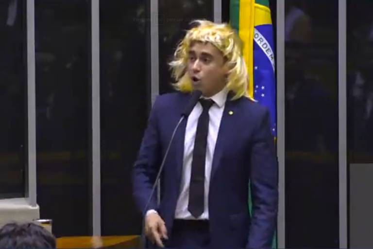 Homem branco de terno e gravata com peruca loira com bandeira do Brasil ao fundo