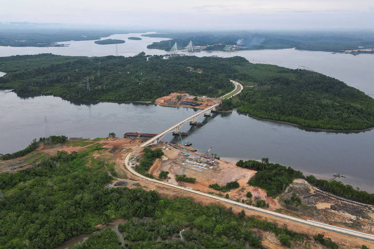 a foto aérea mostra duas porções de terra, com muitas árvores, rodeadas por água. uma ponte em construção conecta os dois espaços