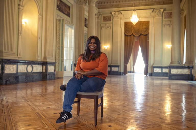 Imagem mostra uma mulher negra jovem sorridente. Ela tem cabelos lisos, usa camisa laranja, calça jeans e tênis adidas preto. Está sentada em uma cadeira em um salão amplo e vazio, com cortinas ao fundo e iluminado por lustre.