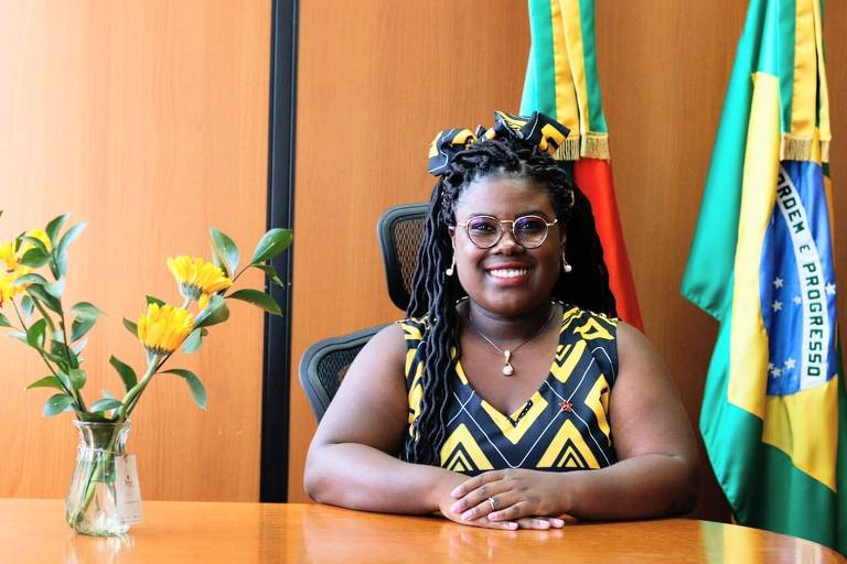 Imagem em primeiro plano mostra mulher negra retinta posando para foto sorrindo. Ela está sentada e apoia as duas mãos em uma mesa