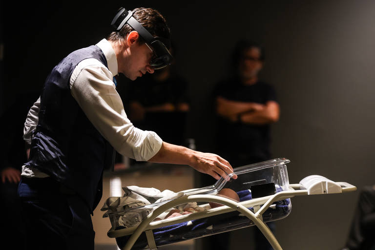 Gabriel Varane, homem branco, veste camisa branca, colete azul e lentes de realidade virtual. À sua frente está um leito de UTI neonatal e um boneco simulador.