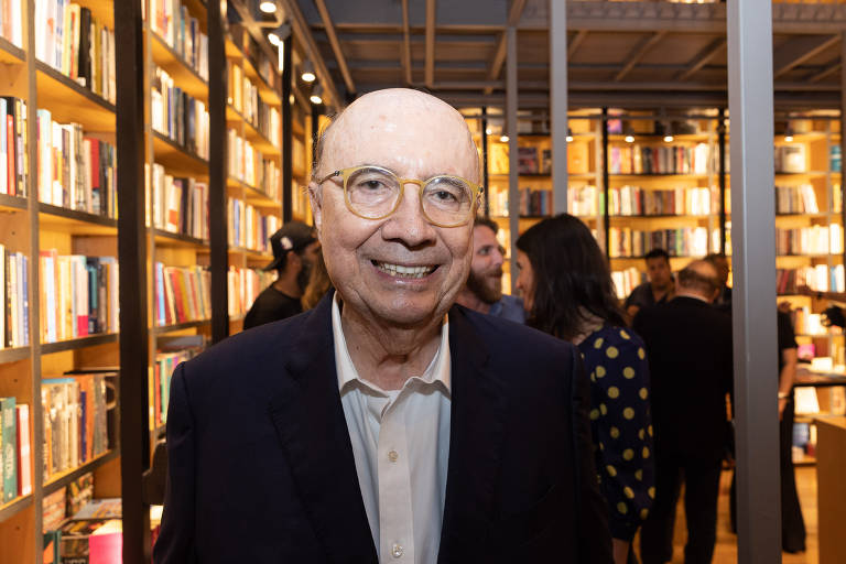 Um homem sorridente está em primeiro plano, posando para a foto em uma livraria. Ao fundo, prateleiras repletas de livros iluminadas por luzes quentes. Há pessoas indistintas ao fundo, criando um ambiente de evento social.