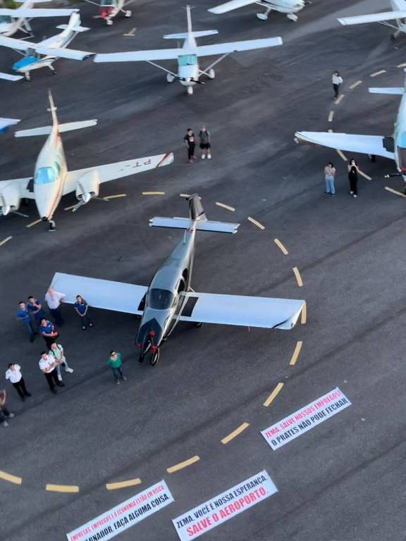 Diversos aviões de pequeno porte na pista de asfalto. Há faixas no chão pedindo ajuda ao governador Romeu Zema