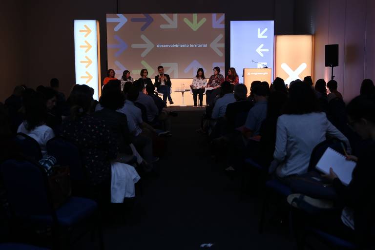 Evento com centenas de pessoas; algumas sentadas em um palco, conversando com uso de microfone, enquanto a plateia ouve