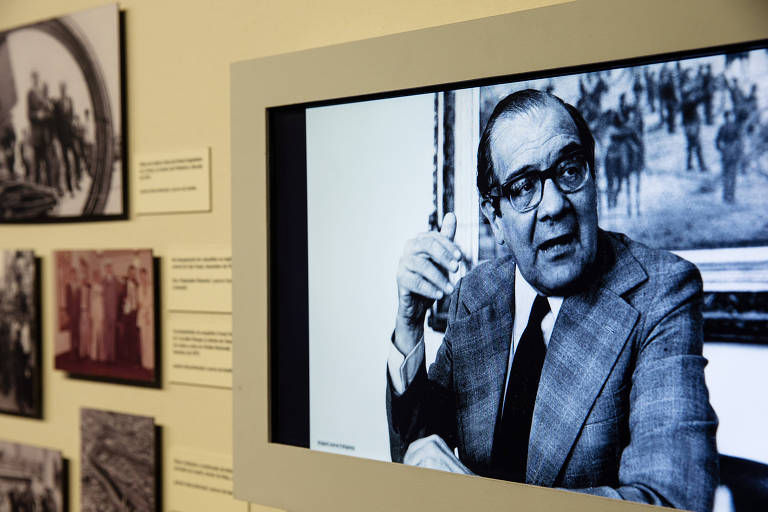 Fotografia de um painel com imagem em preto e branco de homem usando óculos e terno; ao lado, fotografias menores, com textos explicativos
