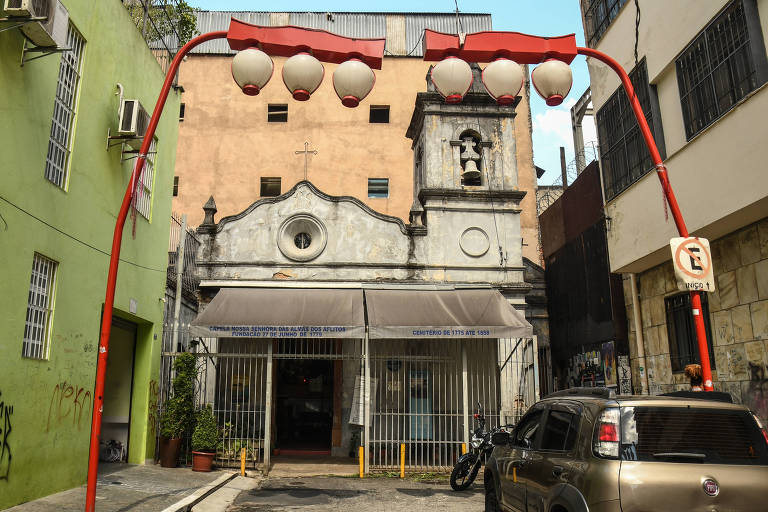 Foto colorida mostra capela branca, pequena, com toldo e gradil de ferro na entrada, atrás de postes em formato de lanternas japonesas. A capela tem uma torre com sino