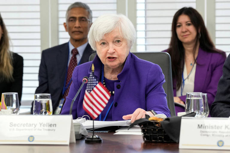 Secretária do Tesouro dos Estados Unidos Janet Yellen participou de mesa redonda, na sede do FMI (Fundo Monetário Internacional), com ministros da economia de outros países. Ao centro da imagem, Yellen é uma mulher branca, de cabelos brancos, vestida em caso violeta. Ela está sentada em uma cadeira de escritório, atrás de uma pequena bandeira dos Estados Unidos.