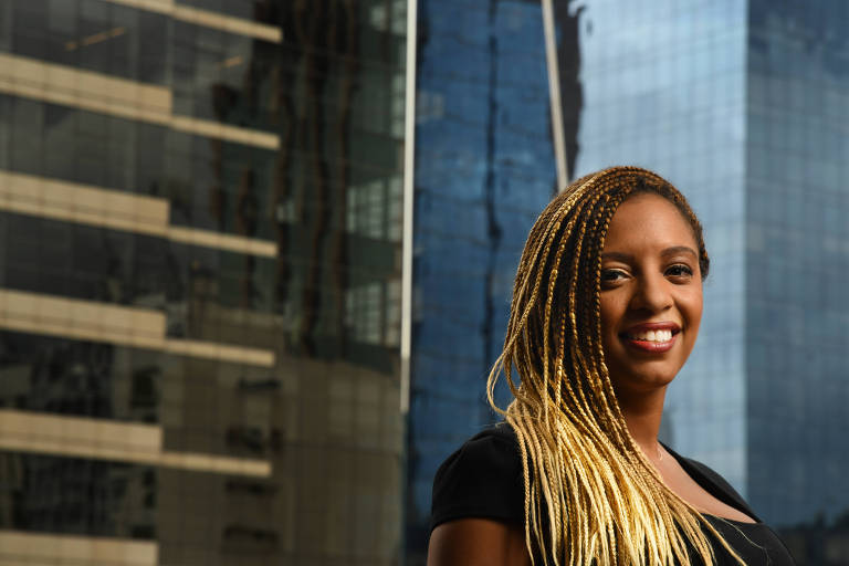 Uma mulher negra sorri diante de prédios com grandes vidraças. Ela sorri, usa uma blusa preta e tranças loiras caídas sobre o ombro direito 