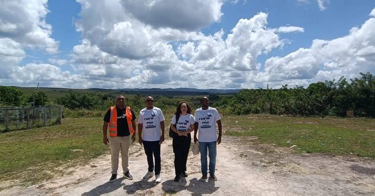 Grupo de quatro pessoas, sendo três homens e uma mulher, está em pé, em uma área aberta com grama e, mais ao fundo, outras plantações. Três deles usam uma camiseta branca da Cufa, ONG brasileira