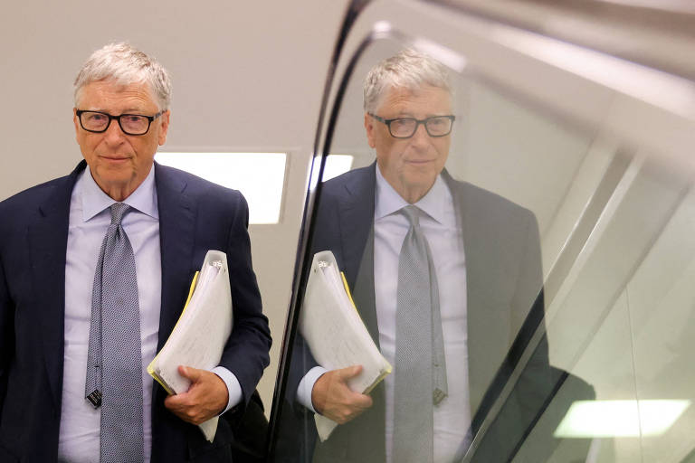 Bill Gates, homem branco, tem cabelos brancos, usa óculos de grau de armação escura. Está de terno e gravata e segurando algo que parece uma pasta ou páginas, branco. Sua imagem está refletida no vidro que aparece na metade direita da foto, vidro da proteção lateral de escada rolante.