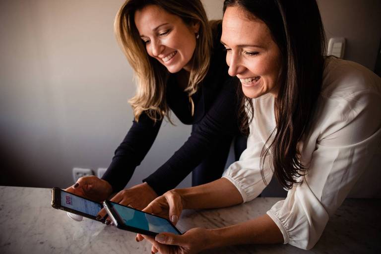 Duas mulheres brancas, lado a lado, apoiam os braços em uma mesa; cada uma segura um celular 