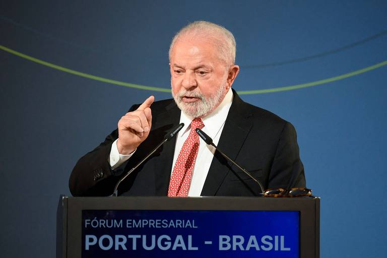 Lula, homem branco, usa camisa branca, gravata em tom vermelho e paletó preto. Ele fala em púlpito onde se lê "Portugal - Brasil" e há dois microfones. Fundo azul com uma fina linha verde