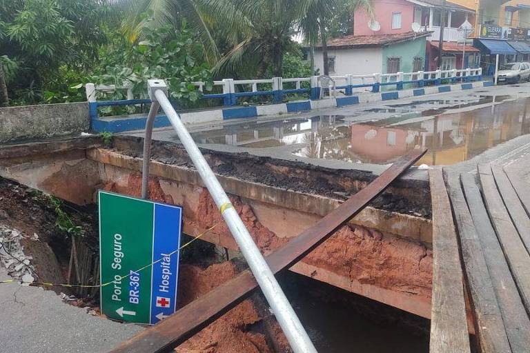 Foto mostra buraco no asfalto na abeceira da ponte, que está interdidata com dosi troncos de madeira cruzados