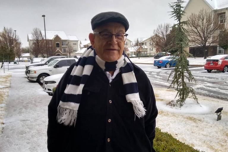 antonio posa para foto, há neve atrás dele na rua e por cima dos automóveis. ele é branco, idoso, sorri, usa boina preta, cachecol listrado preto e branco 