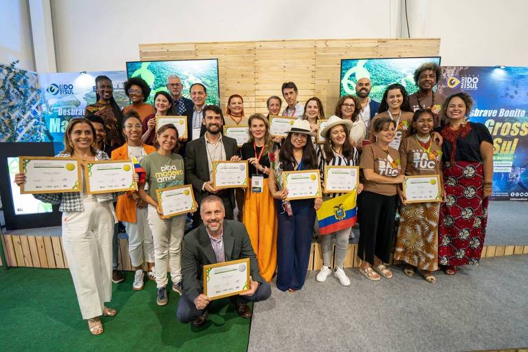 Cerca de 20 pessoas se reúnem à frente de um palco; cada uma segura um tipo de certificado que representa o Prêmio de Turismo Responsável; elas sorriem para a foto e usam roupas formais 