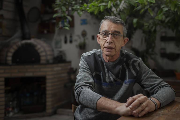 Imagem colorida mostra um homem de 63 anos, branco, de óculos e cabelos grisalhos. Ele veste uma blusa cinza, está sentado, com os braços cruzados em cima de uma mesa.

