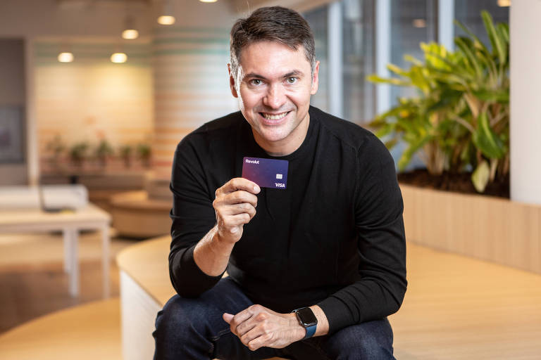 Glauber Mota, CEO da fintech Revolut no Brasil, é um homem branco de cabelos grisalhos com moletom preto sentado em um banco de madeira segurando um cartão com a mão direita à frente do corpo