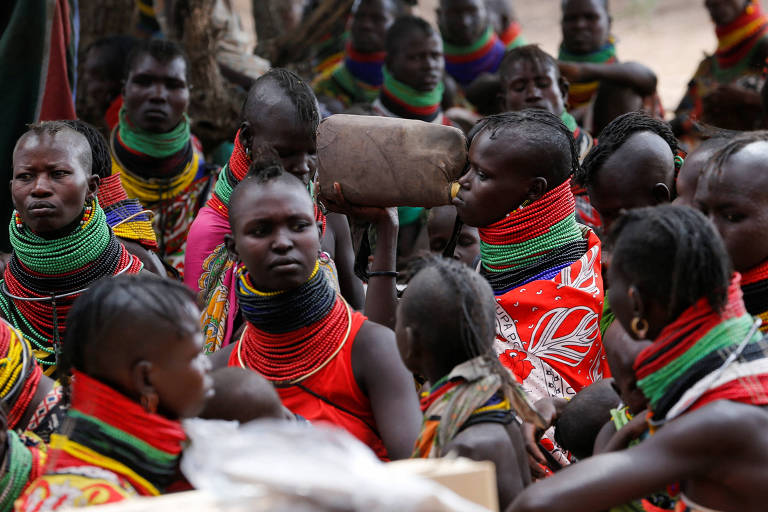 Grupo de mulheres negras com roupas e colares coloridos. Uma delas bebe água de um galão