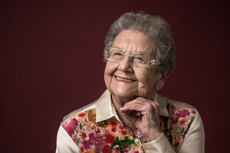 Retrato de Palmirinha, senhora vestida com camisa colorida e mangas bege. Ela leva a mão esquerda sobre o rosto, apoiada ao queixo