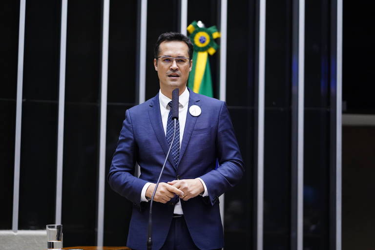 Deltan Dallagnol, homem branco, usa terno azul, camisa branca e gravata. Ele fala ao microfone