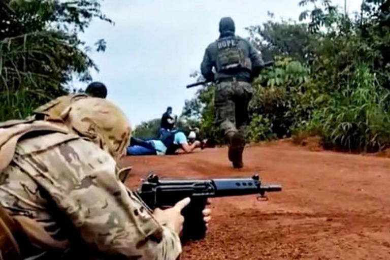 Imagem mostra um homem de traje camuflado bege deitado em uma estrada de chão e apontando um fuzil para a direita, onde se enxerga uma mata. Ao fundo, outro policial de roupa camuflada verde corre na direção contrária à câmera com uma arma na mão.