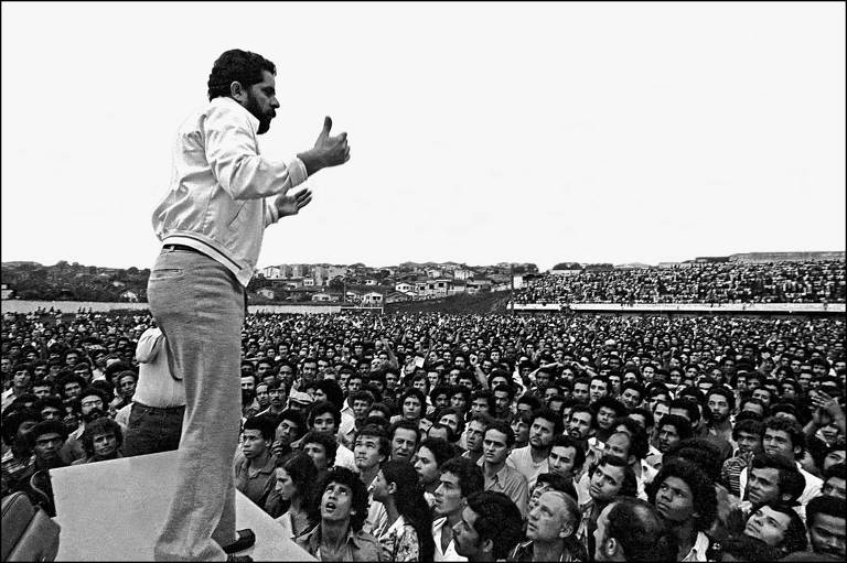 Lula, de calça clara, camisa branca, sobre uma banqueta, gesticula para centenas de operários