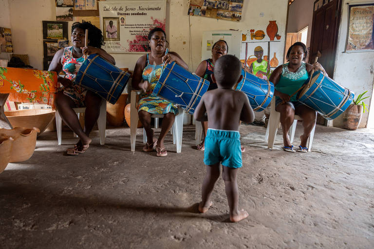 mulheres em roda tocam tambores, em frente a um menino pequeno