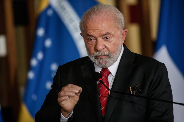 Lula está de pé e fala em ao microfone em coletiva, ele veste terno escuro e gravata vermelha. Ao fundo bandeira do Brasil