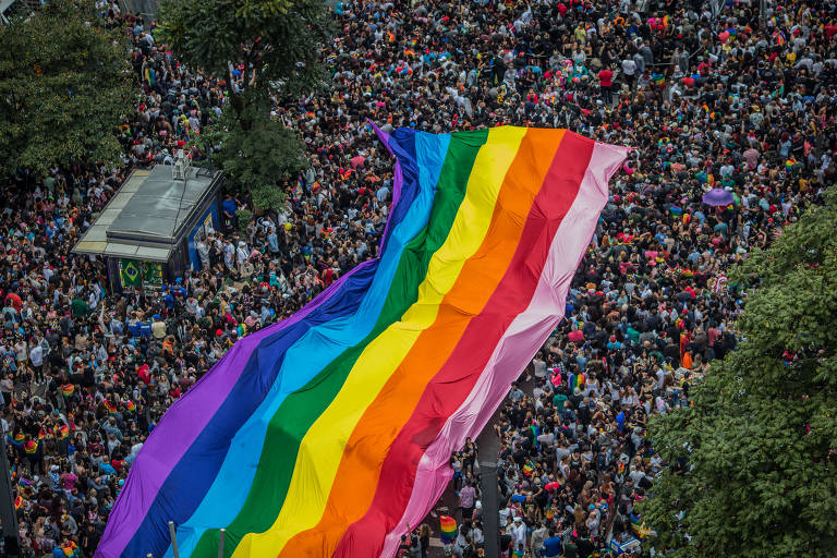 Uma imensa bandeira do arco-íris, símbolo do orgulho LGBTQIA+, ondula acima de uma multidão densa