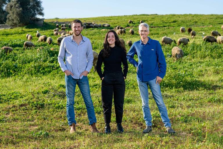 Dois homens e uma mulher posam para retrato em um campo com ovelhas ao fundo