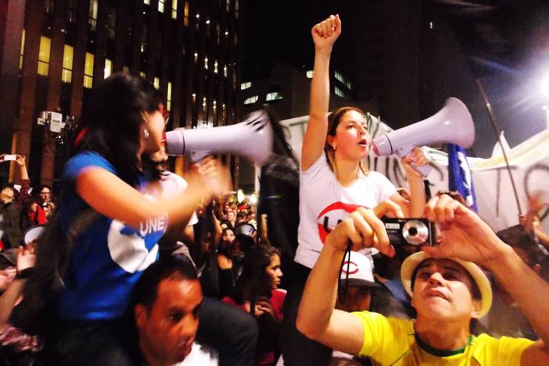 Mulher branca de camiseta branca com nome da UEE, megafone e mão erguida com punho fechado em meio a protesto e outros manifestantes 