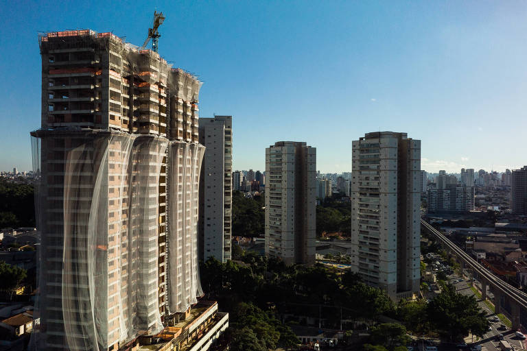 Prédio com mais de 15 anos e três torres em construção, em primeiro plano, com edifícios esparsos entre vegetação e casas no fundo