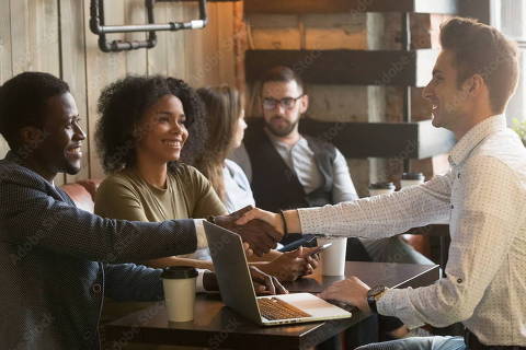 ** IMAGEM EM BAIXA *** Multiracial african and caucasian men handshaking at meeting in cafe, diverse friends greeting sitting at coffeeshop table or black couple making deal shaking hands to white businessman with laptop.Credit Fizkes / Adobe Stock