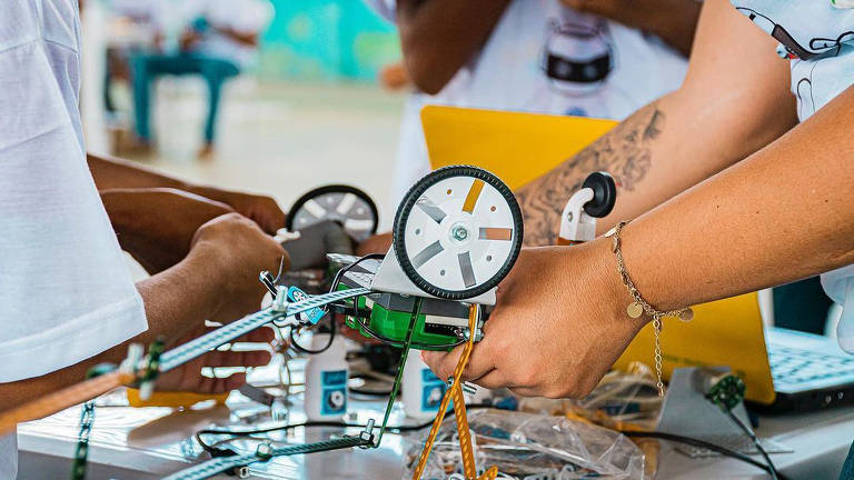 Imagem mostra braços montando um robô verde com rodas brancas e pretas