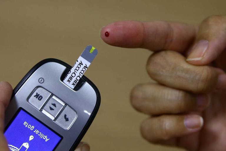 Mão branca com um pouco de sangue no dedo indicador e uma aparelho medidor de insulina ao lado